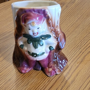 May include: A ceramic planter shaped like a tree trunk with a whimsical elf figure. The elf is wearing a green and white outfit with a brown belt and has a red and white hat. The planter is decorated with brown and white glaze.