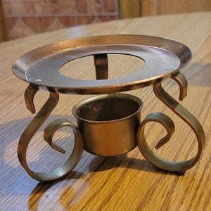 May include: A brass-toned metal tea light candle warmer stand featuring a circular top plate with a central opening, supported by three curved, scroll-shaped legs and a small, cylindrical central cup designed to hold a tea light candle.