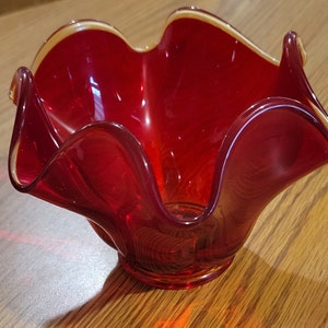 May include: A vibrant red glass bowl with a ruffled rim and a golden-yellow edge. The bowl has a unique, organic shape and sits on a small, circular base. The glass is transparent, allowing light to pass through.