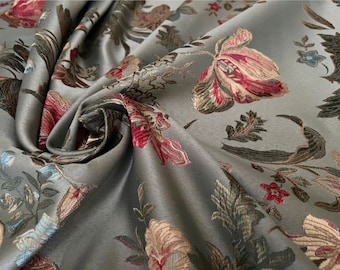 May include: A grey fabric featuring a woven floral pattern with large blossoms in shades of red, pink, and cream, accented by green and dark brown foliage. The material has a smooth, slightly lustrous finish and is draped to show its texture.
