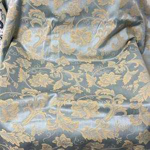 May include: A close-up of a light blue fabric with a floral pattern in a golden-yellow color. The fabric has a slight sheen and is draped over an unseen object. The floral design includes stylized flowers and leaves.