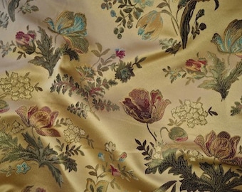 May include: Close-up of a golden fabric with an embroidered floral pattern. The design features large, stylized flowers in shades of burgundy, teal, and cream, along with green foliage. The fabric has a subtle sheen, suggesting a luxurious material.