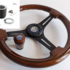 Peut inclure: Un volant en bois brun avec un moyeu central noir et trois rayons noirs. Le moyeu central arbore un logo Alfa Romeo noir avec une croix rouge et un serpent vert. Le volant a un jante en bois brun avec une bande noire.