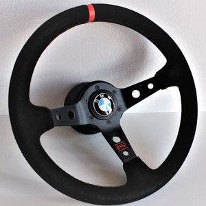 May include: Black suede steering wheel with a red stripe and a black and silver centre hub with a BMW logo. The steering wheel is labelled "OMP".