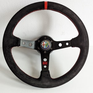 以下が含まれることがあります： 赤いステッチと上部の赤いストライプが付いた黒いスエードステアリングホイール。ホイールは、Alfa RomeoのロゴとOMPのロゴが赤色で配置された黒いセンターを備えています。