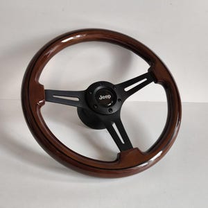 May include: A wooden Jeep steering wheel with a polished finish. The wheel has a dark brown rim and black spokes. The centre features the Jeep logo in white on a black background.
