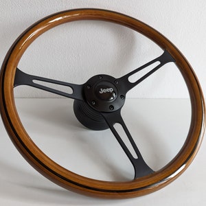 May include: A wooden Jeep steering wheel with a polished finish. The wheel features a three-spoke design with black metal spokes and a black centre hub with the Jeep logo. The wood is a warm brown colour.