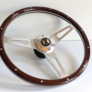 Peut inclure: Un volant en bois brun avec une jante argentée et trois rayons argentés. Le volant a un capuchon central argenté avec un logo Volkswagen.