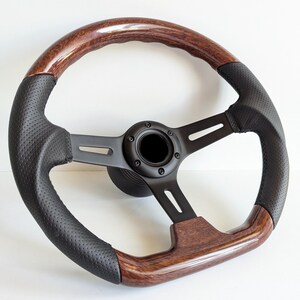 Può includere: Un volante per auto classico con un bordo in legno scuro e un'impugnatura in pelle nera traforata. Il volante ha un telaio e un mozzo centrale in metallo nero. Le venature del legno hanno una finitura lucida.