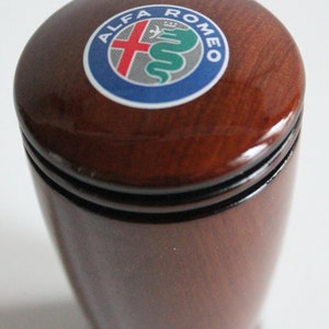 May include: A brown wooden gear shift knob with a black ring around the top and a silver, red, and green Alfa Romeo logo.