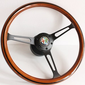 Könnte beinhalten: Ein klassisches Holzlenkrad mit dunkelbraunem Finish und schwarzen Akzenten. Das Lenkrad hat ein dreispeichiges Design mit einem schwarzen Mittelteil, das das Alfa Romeo-Logo zeigt. Der Felgenrand hat eine glänzende Oberfläche mit einem dünnen schwarzen Streifen.