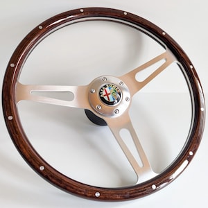 Pode incluir: Um volante de madeira escura com raios de metal escovado. O centro apresenta um emblema da Alfa Romeo. O volante é adornado com rebites prateados. O diâmetro é de aproximadamente 38 cm.