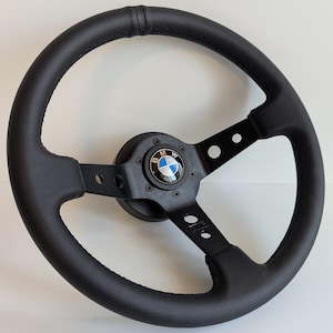Könnte beinhalten: Schwarzes Lederlenkrad mit Dreispeichen-Design. In der Mitte befindet sich ein BMW-Logo in Blau, Weiß und Schwarz. Das Lenkrad hat einen genähten Griff und eine mattschwarze Oberfläche.