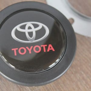 Puede incluir: Emblema Toyota negro y plateado. El logotipo de Toyota es plateado y la palabra "TOYOTA" es roja. El emblema es redondo con un borde negro. Un anillo plateado es visible en el fondo.