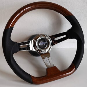 Può includere: Un volante in legno marrone con impugnatura in pelle nera e un centro cromato con un logo Ford blu.