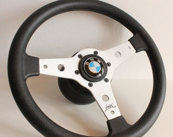 May include: A black leather steering wheel with a silver center hub and a blue and white BMW logo. The steering wheel has three spokes and a black leather grip.