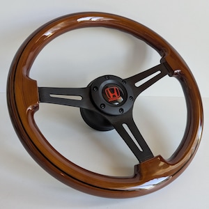 Può includere: Un volante in legno con finitura lucida. Il volante ha un design a tre razze con razze in metallo nero e un emblema centrale con il logo Honda in rosso e bianco.
