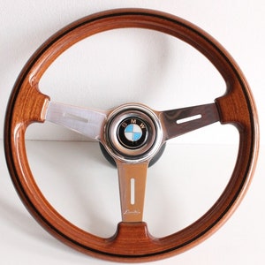 Può includere: Un volante BMW classico in legno. Il volante presenta un design a tre razze con un centro argentato e l'emblema BMW. Il legno è marrone caldo con dettagli neri.