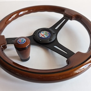 Pode incluir: Um volante de madeira castanho com um cubo central preto e três raios pretos. O volante tem uma moldura preta em torno da borda. Uma alavanca de mudanças de madeira castanho com um logotipo Alfa Romeo prateado está presa ao volante.