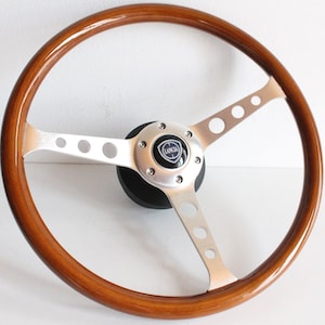 May include: A brown wooden steering wheel with a silver spoke design and a silver centre hub with a blue and silver Lancia logo.
