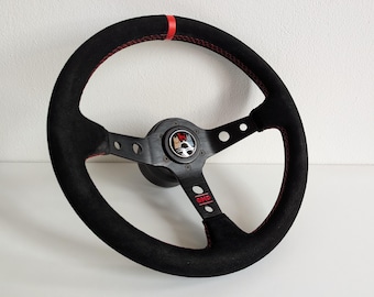 May include: Black suede steering wheel with red stitching and a black center hub with a silver logo. The steering wheel has three spokes and a red "OMP" logo.