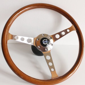 Könnte beinhalten: Ein klassisches Holzlenkrad mit polierter Oberfläche. Das Lenkrad hat ein dreispeichiges Design mit Chromakzenten und einem Mittelteil mit dem Volvo-Logo. Der Durchmesser des Lenkrads beträgt ca. 35,5 cm.