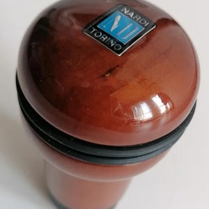 May include: A brown wooden gear shift knob with a black rubber ring around the base. The knob has a blue and silver Nardi Torino logo on the top.