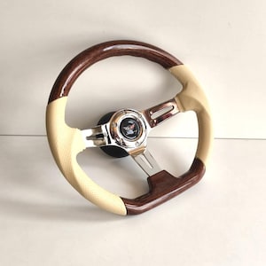 Pode incluir: Um volante de carro clássico com uma jante de madeira e uma pega em couro perfurado creme. O centro apresenta um emblema cromado com fundo preto. Os raios são cromados.