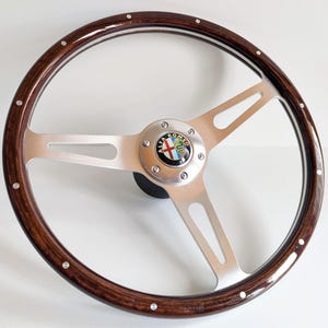 Può includere: Un volante in legno classico con finitura a venature scure. Presenta razze in metallo spazzolato e un emblema centrale con la scritta "ALFA ROMEO". Rivetti argentati accentuano il volante. Diametro di circa 38 cm.