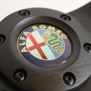 Puede incluir: Volante negro con un logotipo Alfa Romeo plateado en el centro. El logotipo tiene una cruz roja, una serpiente verde y las palabras "Alfa Romeo" en un c&iacute;rculo blanco.