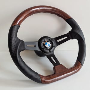 May include: A black and wood-grain steering wheel with a BMW logo in the center. The wheel has a black perforated leather grip and a dark wood-grain finish on the top and bottom. The spokes are black.