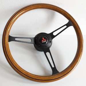 May include: A brown wood steering wheel with a black leather rim and three black spokes. The center of the steering wheel has a black hub with a silver Mitsubishi logo.