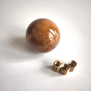 May include: A spherical wooden shift knob with a polished, glossy finish, displaying a warm brown hue and wood grain. Three brass threaded inserts are also present. The knob is likely for a vehicle.