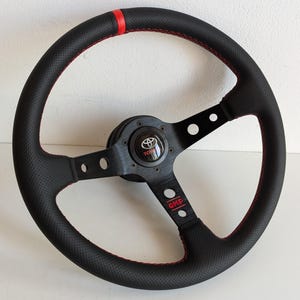 May include: Black perforated leather steering wheel with red stitching and a red stripe. Features a black centre with the Toyota logo and the OMP logo. The wheel has a three-spoke design.