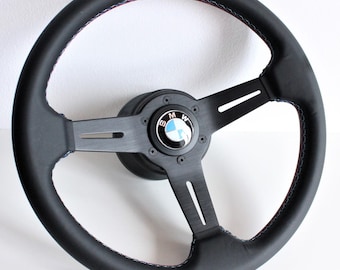 May include: Black leather steering wheel with a three-spoke design and a blue and white BMW logo in the center. The wheel has red and blue stitching around the rim.
