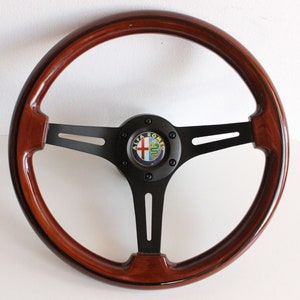 Puede incluir: Un volante de madera marr&oacute;n con un centro negro y un logotipo de Alfa Romeo negro. El volante tiene tres radios.