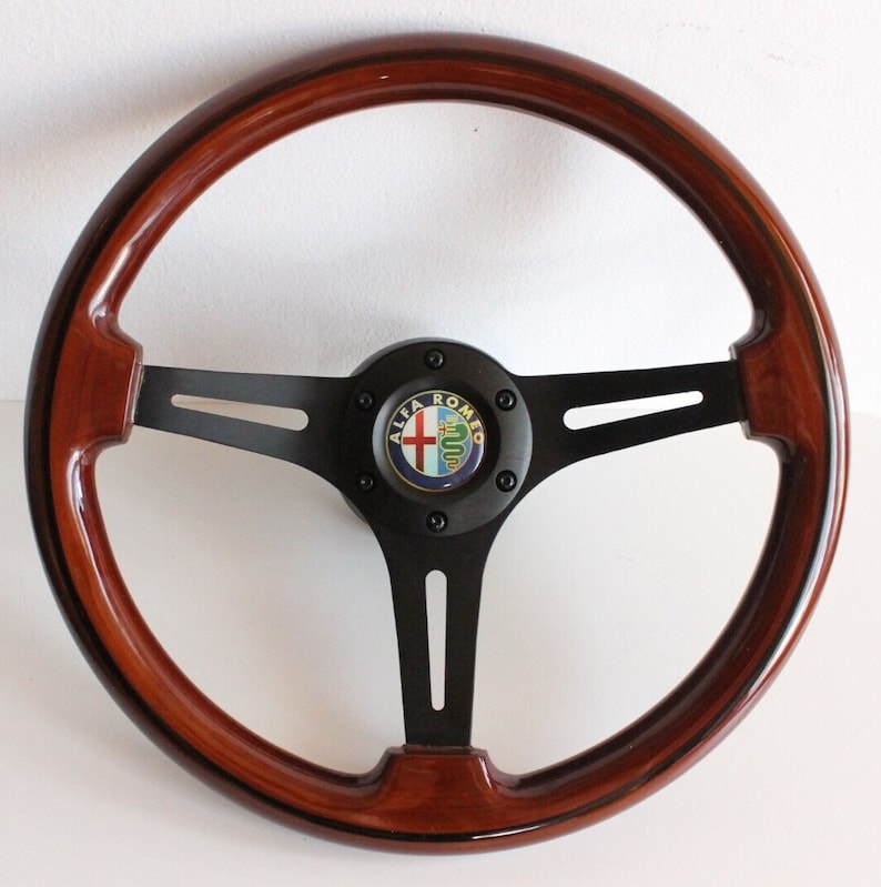 Puede incluir: Un volante de madera marr&oacute;n con un centro negro y un logotipo de Alfa Romeo negro. El volante tiene tres radios.