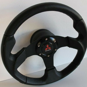 Può includere: Volante in pelle nera con un mozzo centrale nero e argento con il logo Mitsubishi in rosso.