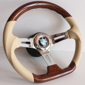 Puede incluir: Un volante de madera marrón con un borde de cuero beige y un centro cromado con el logotipo de BMW. El volante tiene tres radios.