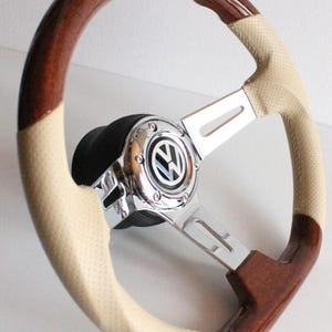 Puede incluir: Un volante de coche clásico con un borde de madera pulida y un agarre de cuero perforado de color crema. El centro presenta un emblema cromado con el logotipo de Volkswagen. El volante tiene un acabado brillante y una estética vintage.