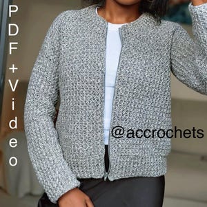 Op de afbeelding: Een grijze, getextureerde gehaakte jas met ritssluiting. De jas heeft lange mouwen en een ronde halslijn. De tekst "PDF + Video" staat links en "@accrochets" rechts in de afbeelding.