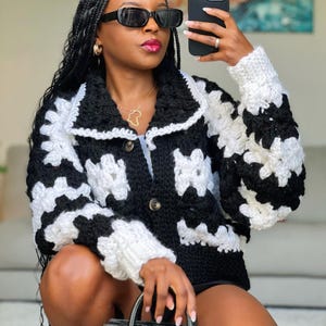 May include: A black and white crochet cardigan with a button-up front. The jacket has a geometric pattern and a collar. The sleeves are long and the cardigan is worn with sunglasses and a black handbag.