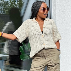 May include: A white crochet top with a button-up front and short sleeves. The top is worn over a black vest top and paired with khaki cargo trousers.