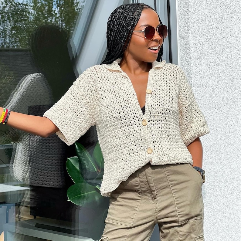 May include: A white crochet top with a button-up front and short sleeves. The top is worn over a black vest top and paired with khaki cargo trousers.