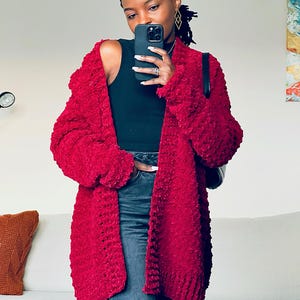 Op de afbeelding: Een lange, rode cardigan met een pluche textuur. De cardigan is open en de persoon die hem draagt ​​houdt een telefoon vast.