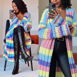 May include: A long, open-front crochet cardigan in pastel shades of blue, yellow, pink, and purple. The cardigan features long sleeves and a V-neck. The garment is worn with black trousers and boots. The crochet pattern is visible.