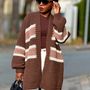 Peut inclure: Un cardigan marron à devant ouvert avec des rayures blanches et roses. Le pull en tricot épais à manches longues est porté avec un pantalon blanc et un sac à main noir. La personne porte des lunettes de soleil et un foulard noir.
