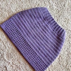 Peut inclure: Un col en crochet violet fait à la main. Le col présente un motif côtelé avec une bordure texturée. L'article est fait de fil doux et convient aux temps plus frais. Le col est présenté sur un tapis de couleur neutre.