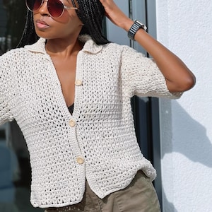 Puede incluir: Una camisa blanca de crochet con botones y mangas cortas. La camisa tiene un corte holgado y está hecha con un hilo ligero.