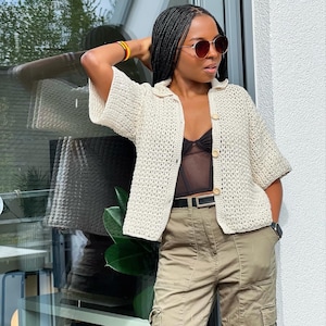 May include: A white crochet cardigan with a button fastening and a black lace bralette underneath. The cardigan has a loose fit and short sleeves. The model is wearing khaki cargo trousers and sunglasses.